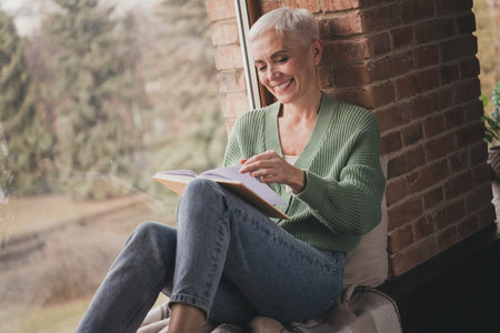 Elderly woman enjoying a cozy day indoors while reading and smiling in stylish living room settingの写真素材