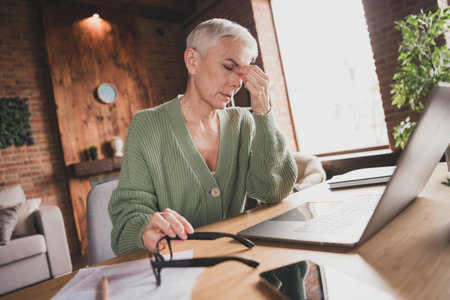 Elderly woman in stylish green cardigan working on laptop, feeling tired at home in bright living roomの写真素材