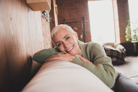 Charming senior woman in cozy green sweater relaxing at home with a happy smile, enjoying a peaceful...