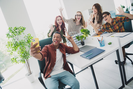 Photo of smiling happy web designers working startup together showing v-signs recording video gadget...