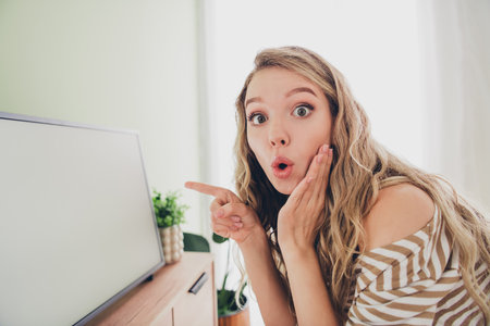 Surprised young woman with blond hair pointing at blank screen, in cozy living room setting, wearing striped pullover, capturing a moment of youthful charm and curiosity.の写真素材