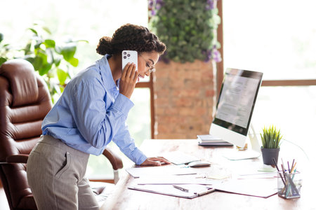 Focused businesswoman on a call at her desk, multitasking confidently in a bright modern officeの写真素材