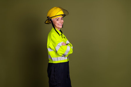 Confident mature woman in high-visibility workwear and safety helmet, standing with arms crossed, isolated against khaki backgroundの写真素材