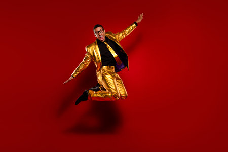 Dynamic young man in a shiny golden suit jumping joyfully against a bold red backgroundの写真素材