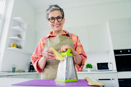 Charming mature woman in apron grating cheese in bright modern kitchen, showcasing skilled cooking and culinary expertiseの写真素材