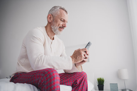 Elderly man in pajamas sitting on bed using smartphone in bright bedroom during peaceful morningの写真素材