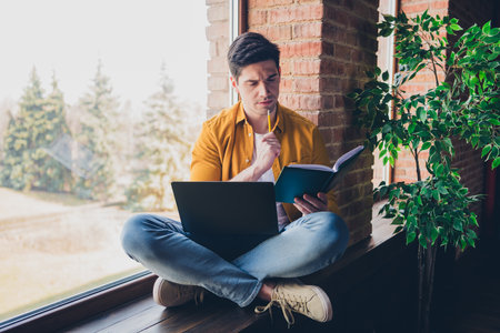 Young man relaxing at home, seated by window with laptop and book, enjoying daylight