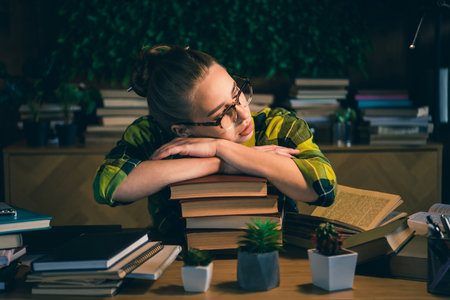 Young woman with blond hair in checkered shirt studying at home, leaning on books, and feeling relaxed in cozy indoorの写真素材