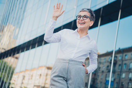 Confident mature woman with short grey hair waves in urban cityscape, wearing elegant blouse and formalwearの写真素材