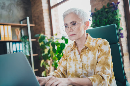 Photo of lovely pensioner lady typing netbook working dressed yellow print shirt formalwear comfortable startup office loft room interiorの写真素材