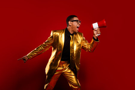 Vibrant young man in a glossy golden suit joyfully holds a red megaphone against an elegant red...