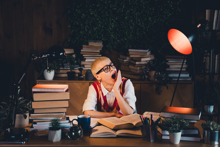 Young woman yawning in dimly lit study room, surrounded by stacks of books, wearing glasses and a casual vest over a white shirtの写真素材