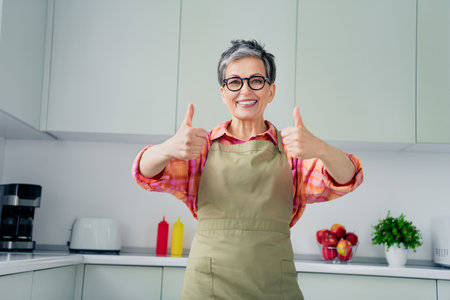 Charming elderly woman in apron gives thumbs up in a modern kitchen with daylight streaming in, showcasing comfort and joy in cookingの写真素材