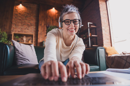 Happy elderly woman with short grey hair in casual style, enjoying leisure time indoors, typing on laptop with a cheerful smileの写真素材