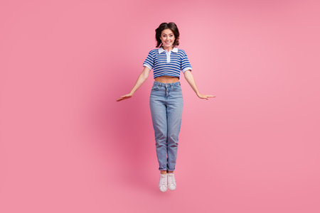 Young woman in casual striped t-shirt and jeans jumping joyfully against pink background, exuding...