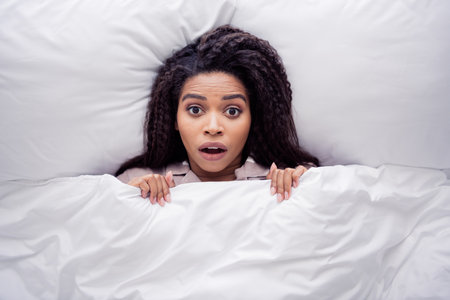 Surprised young woman in sleepwear waking up in cozy bedroom with natural daylightの写真素材