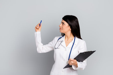 Young female doctor in white coat holding clipboard and pen against grey backgroundの写真素材