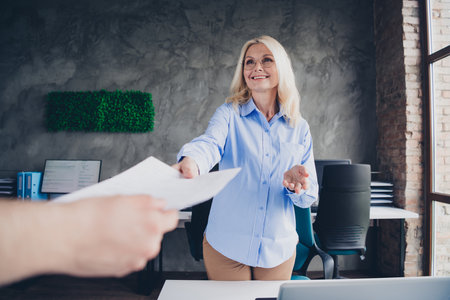 Mature businesswoman handing document while working in modern office with natural daylightの写真素材