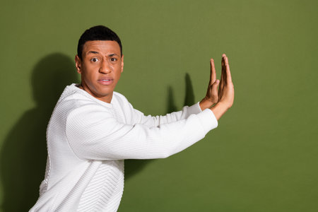 Young handsome man showing stop hand gesture on khaki background, wearing stylish white sweater, expresses mixed emotionsの写真素材