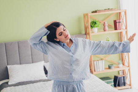 Charming young woman in blue sleepwear stretching happily while awakening on a sunny morning in her cozy bedroomの写真素材