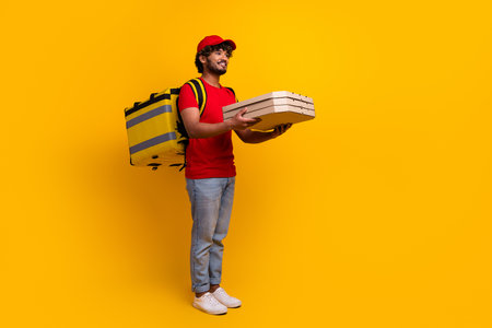 Young courier wearing red uniform holding pizza boxes while standing with yellow thermal bag against yellow backgroundの写真素材