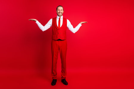 Full size photo of handsome young guy hands compare hold empty space wear trendy classic outfit isolated on red color backgroundの写真素材