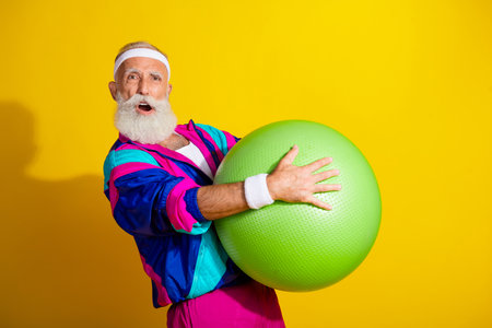 Photo of handsome excited senior guy oldschool sportswear enjoy fitball exercises emtpy space isolated yellow color backgroundの写真素材