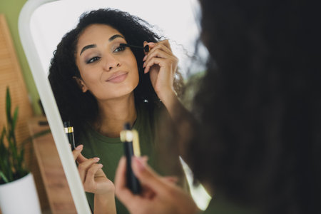 Photo of attractive young woman apply mascara mirror reflection dressed green outfit daylight home...
