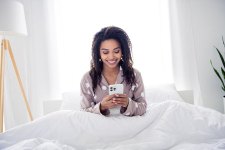 Young woman enjoying a morning text on her phone in a cozy bedroom environment with bright natural lightの写真素材