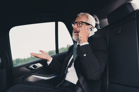 Mature businessman in a suit engaged in an animated phone conversation while traveling in a car on a...
