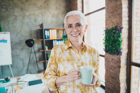 Photo of pretty retired female hold coffee mug wear yellow print shirt formalwear coworking successful nice light officeの写真素材
