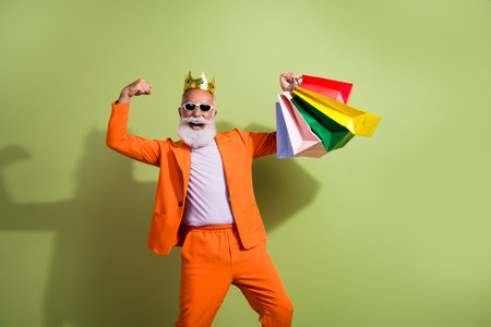 Photo of funny extravagant man golden crown raise fist hold bags wear orange suit isolated on green color backgroundの写真素材