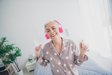 Senior woman in polka dot pajamas enjoys music with pink headphones in her bedroomの写真素材