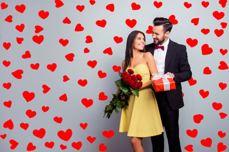 Portrait with copy space of attractive couple in formal wear, woman in dress holding bouquet of red roses, man in tux embracing her from back side and presenting a packaging red case with giftの写真素材