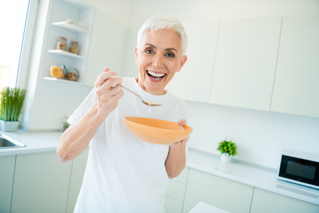 Happy middle-aged woman with short hair enjoys cooking in a bright kitchen, holding a bowl and spoonの写真素材