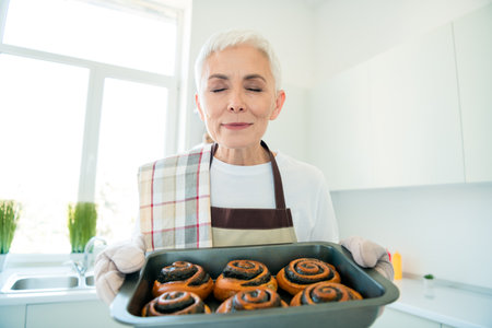 Charming elderly woman enjoying freshly baked cinnamon rolls in her cozy kitchen, with warm sunlight streaming in through the windowの写真素材