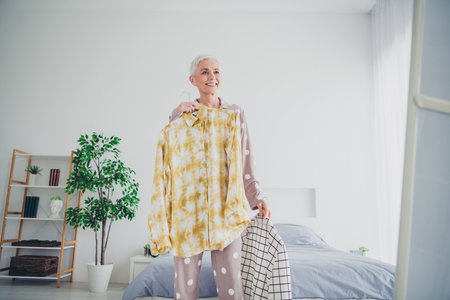Elegant elderly woman in cozy pajamas stands cheerfully in a bright bedroomの写真素材