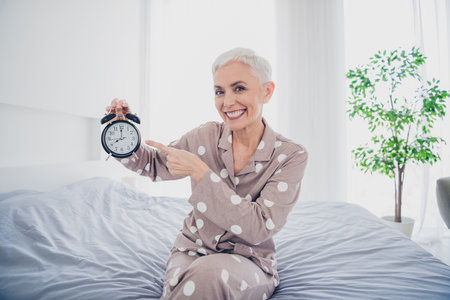 Cheerful senior woman in polka dot pajamas holds alarm clock on a cozy morning at home