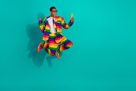 Charismatic young man in a colorful rainbow suit jumping against vibrant turquoise backgroundの写真素材