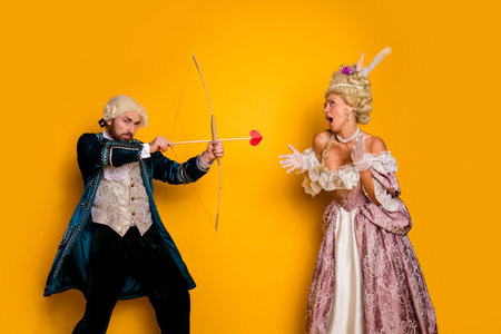 Couple in historical costumes with bow and love arrow against vibrant yellow background depicting...