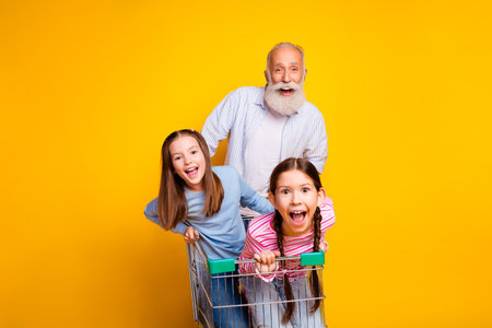 Joyful elderly man pushing two excited girls in a shopping cart against a yellow backdrop with fun expressionsの写真素材