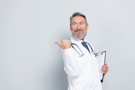 Experienced male doctor in white medical uniform holding clipboard against light grey background
