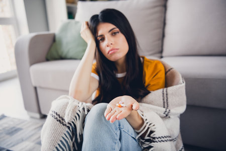 Young woman indoors holding pills, appearing thoughtful in casual settingの写真素材