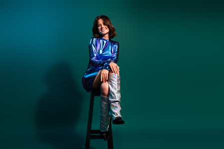Full body portrait of stunning young woman sit chair empty space wear shiny dress neon light dark blue backgroundの写真素材