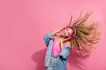 Stylish young woman with headphones and denim jacket enjoying music against pink backgroundの写真素材