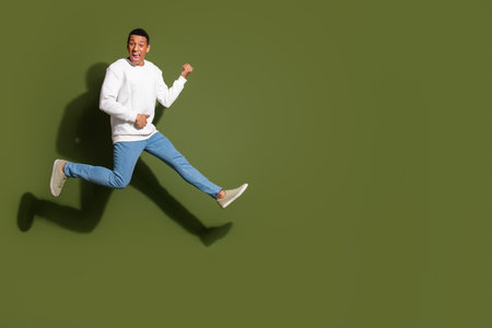 Cheerful young man in a white sweater and jeans leaps joyfully in front of a khaki background, displaying a vibrant expressionの写真素材