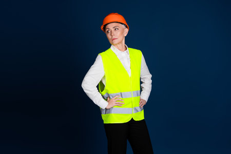 Confident mature female construction worker in uniform and helmet against a dark blue background.