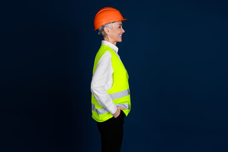 Profile of a mature female construction worker wearing a hardhat and high-visibility vest against a dark blue background.の写真素材