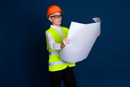 Mature female construction professional in uniform, inspecting blueprints against a dark blue backgroundの写真素材