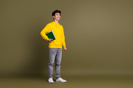 Happy student in casual yellow hoodie holding books standing on khaki background, smiling confidentlyの写真素材